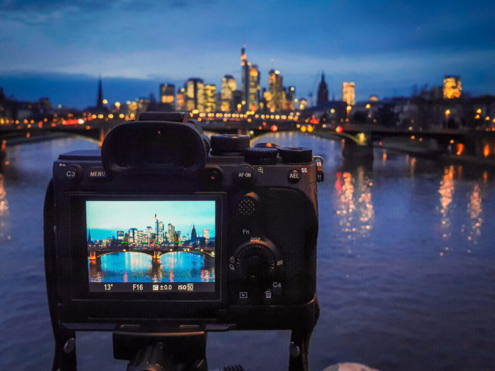 A full frame mirrorless camera being used on a tripod to capture a long exposure image of Frankfurt's cityscape, with the reflections in the river. The device screen shows the settings to be used - a 13 second exposure at F16, with an ISO of 50.