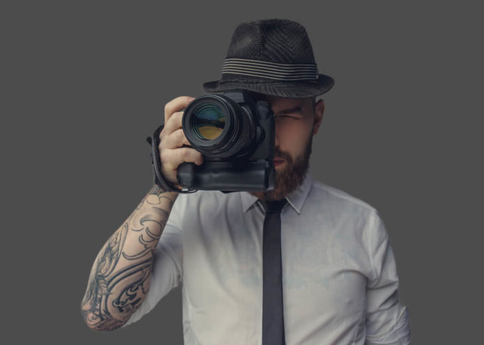 Digital photographer with an expensive digital camera in white shirt and casual hat. Isolated on grey background.